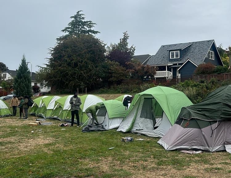 Green tents on a lawn with houses in the background. some people stand near the tents